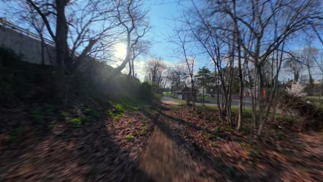 Uma tomada FPV de baixa altitude de uma trilha de terra ladeada por árvores sem folhas no Elmont Road Park em uma manhã ensolarada. A câmera se move lentamente sobre o caminho, rente ao chão