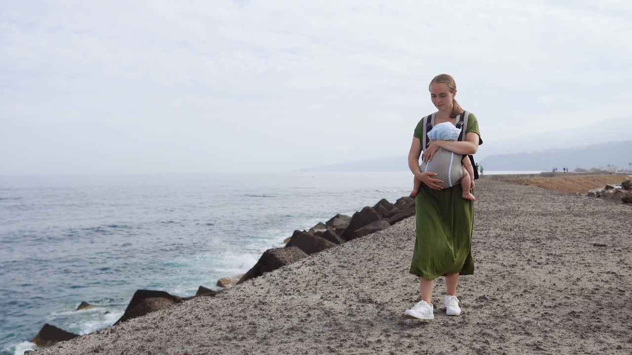 In the embrace of a summer sea, a mother and her baby relish a leisurely walk outdoors. The child is snugly carried in a kangaroo backpack