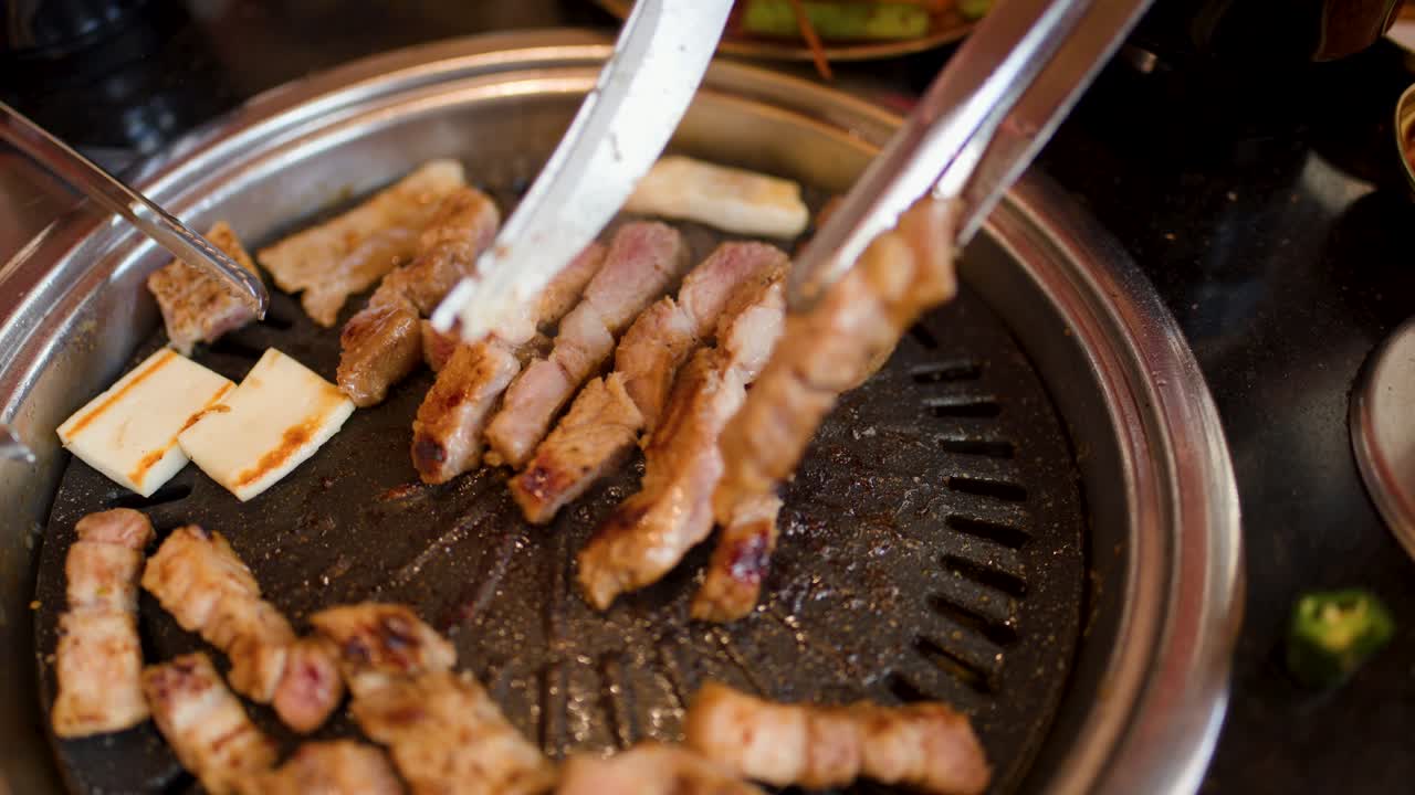 Close-up of pork belly grilling on tabletop barbecue with tongs, shallow focus, warm lighting