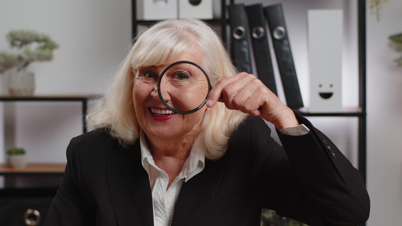 Una donna d'affari anziana sorridente che guarda attraverso la lente d'ingrandimento con un'espressione "wow" in ufficio a casa