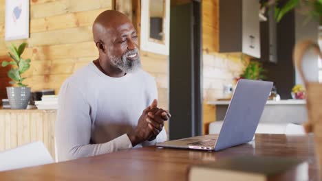 Happy-senior-african-american-man-in-log-cabin,-using-laptop-for-video-call,-slow-motion