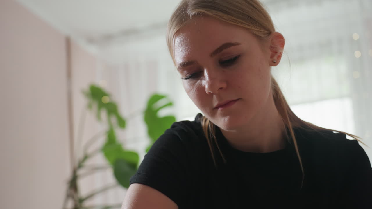 Pretty blonde woman in black shirt with hair tied back looking down with focused expression, softly lit background featuring light-colored wall, large green plant, and window with sheer curtains