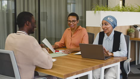 an woman and a muslim woman co workers interview a young man sitting at a table in the office 3