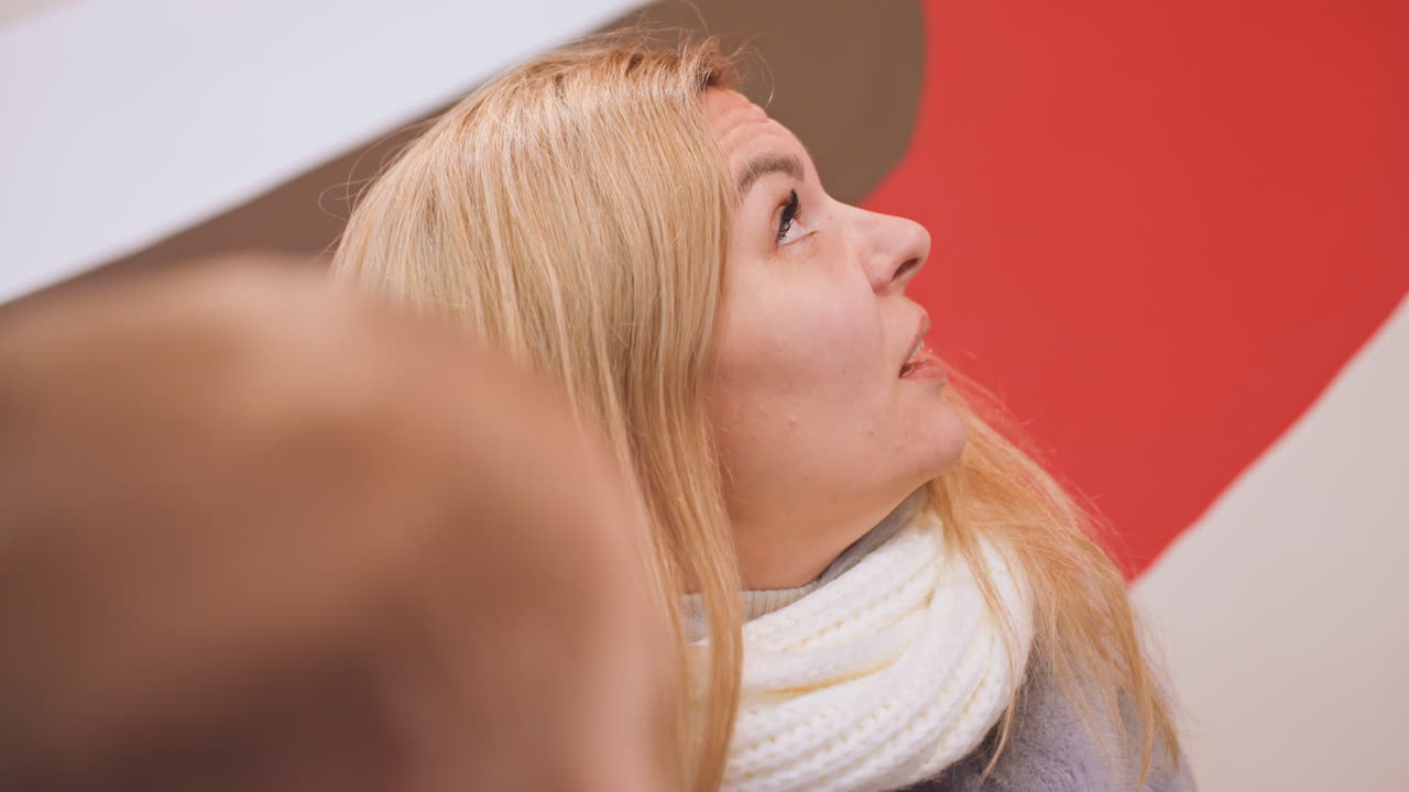 Mama und Sohn im Café, blonde Mutter mit weißem Schal spricht mit kleinem Jungen über einem Getränk. Rotes Wandbild und kleines Regal mit Sukkulente schaffen warme Innenatmosphäre, authentische Ausdrücke und aufmerksamer Blick eingefangen.