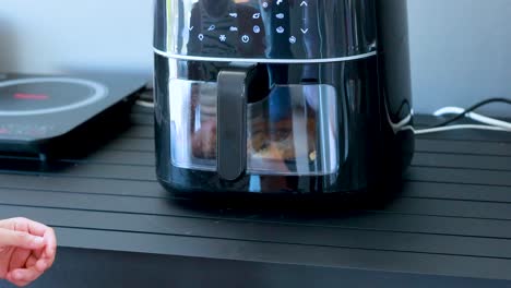 a person activates a digital air fryer on a kitchen counter, pressing the power button with a thumbs-up gesture in bright, natural lighting