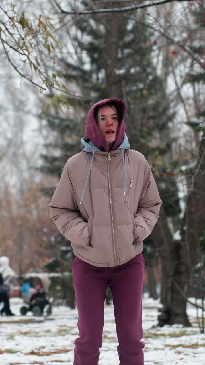 어린 소녀는 따뜻한 후디와 코트를 입고 겨울 스노우 파크에서 놀이하게 뛰어내립니다. 눈으로 인 나무들이 그녀를 둘러싸고 있고, 배경에 있는 사람들은 베이비 워커를 밀고 있습니다.