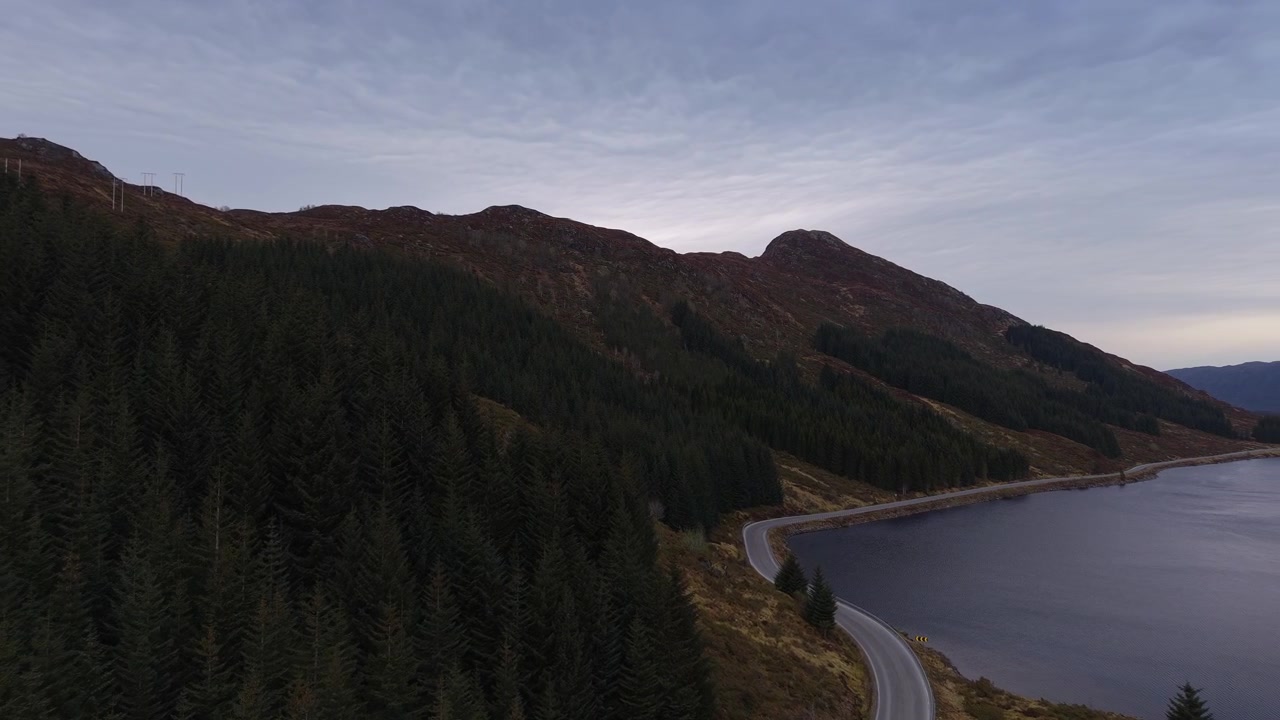 Una strada tortuosa costeggia un lago, fiancheggiata da fitte foreste e colline scoscese sotto un cielo crepuscolare. Ideale per servizi di viaggio o documentari paesaggistici.