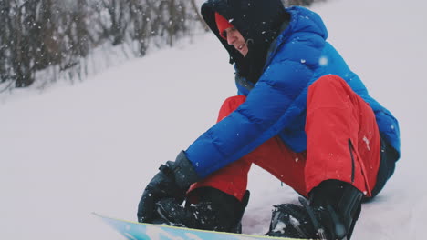 a man in red pants sitting on the snow fastens snowboard shoes and a blue jacket on the ski slope