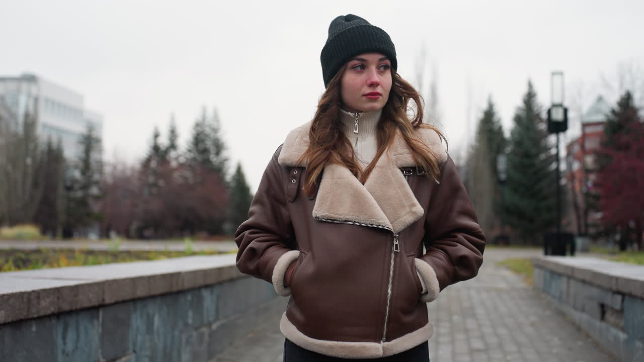 Portrait shot of lady in black cap brown shearling jacket walking slowly with hands in pockets on wide stone pathway through quiet park lined with pine trees modern buildings and cloudy autumn sky