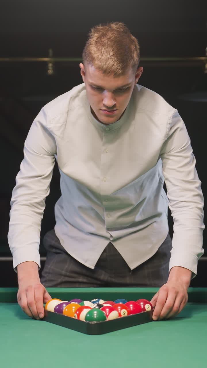 jugador de billar con camisa blanca y pantalones grises arregla cuidadosamente bolas de billar de colores en formación de triángulo en una mesa de billar verde vibrante bajo una cálida iluminación ambiental