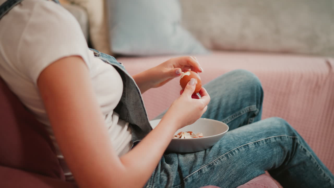 Side view of young girl seated on soft chair wearing denim overalls peeling egg shell slowly and carefully into bowl, with cozy background showing neutral-toned sofa and light blue pillow