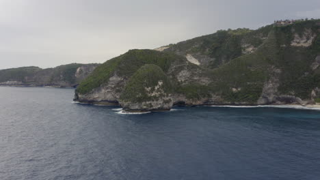 Fly-by-along-the-steep-coast-line-of-Nusa-Penida-with-remote-islands-and-ocean-waves-crushing-ashore,-Bali,-Indonesia