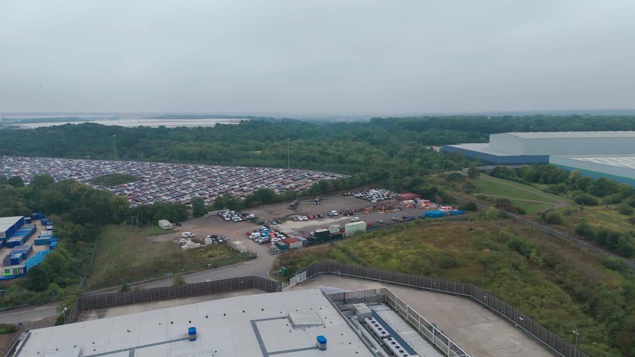 An aerial shot captures an industrial landscape with buildings and a vast parking lot filled with cars. The surrounding greenery and a river in the distance under a cloudy sky add depth to the scene