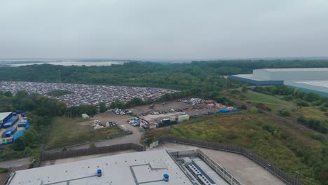 an aerial shot captures an industrial landscape with buildings and a vast parking lot filled with cars. the surrounding greenery and a river in the distance under a cloudy sky add depth to the scene