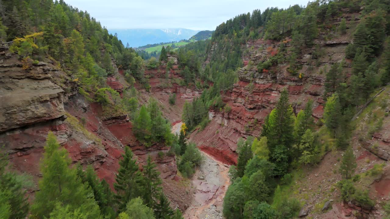 An aerial view of Bletterbach Gorge in South Tyrol, Italy, showcasing impressive layered red rock formations and dense forest. This scenic canyon landscape reveals unique geological features.
