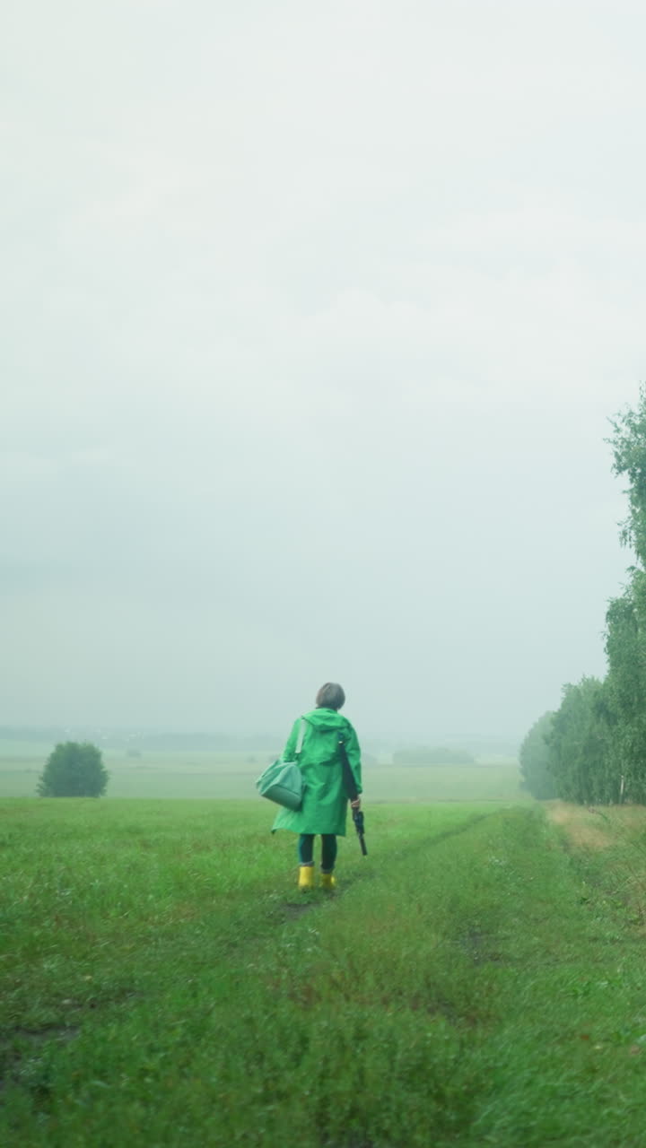 초록색 레인코트를 입고 민트색 가방과 우산을 들고, 노란색 레인 부츠를 입고, 높은 나무, 초록색, 안개 하늘에 둘러싸인 멀리 있는 진길을 고 있는 여성의 뒷면