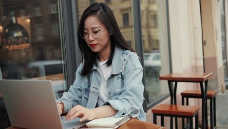 retrato de la atractiva joven con anteojos trabajando en la computadora portátil en la terraza exterior del café y sonriendo a la cámara con alegría