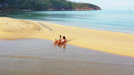 beautiful young women in swimsuit relaxing on sandy stripe beach washed by calm clear water of shallow lagoon on shore of tropical island in bali