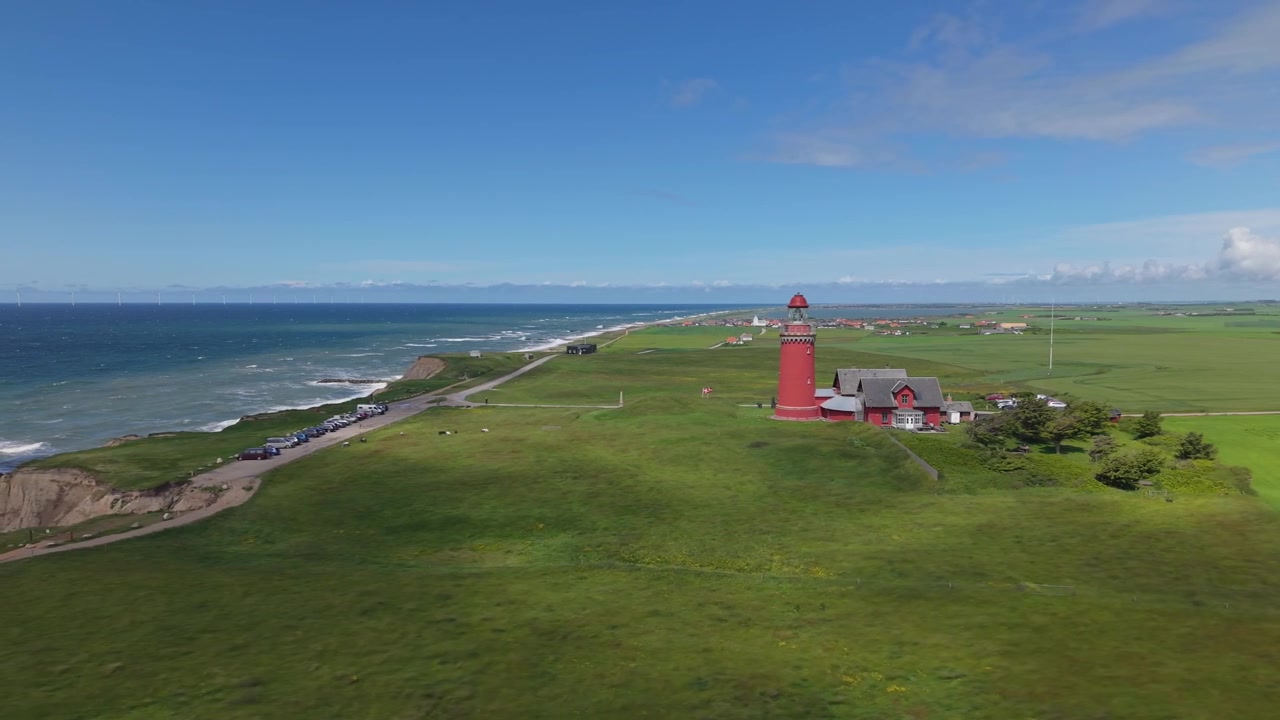 Flygfoto från sidan som fångar det röda Bovbjerg fyr, omgiven av gröna ängar medan rörliga molnskuggor sveper över fälten, med det blå Nordsjön i bakgrunden.