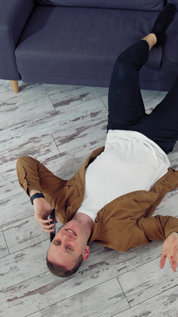 Young man speaking the phone at home. Tired man working through the phone while lying on a floor. Top view. Vertical video
