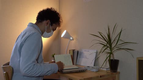 man wearing a face mask sitting while reviewing his office notes during a home quarantine due to covid-19 outbreak - medium shot