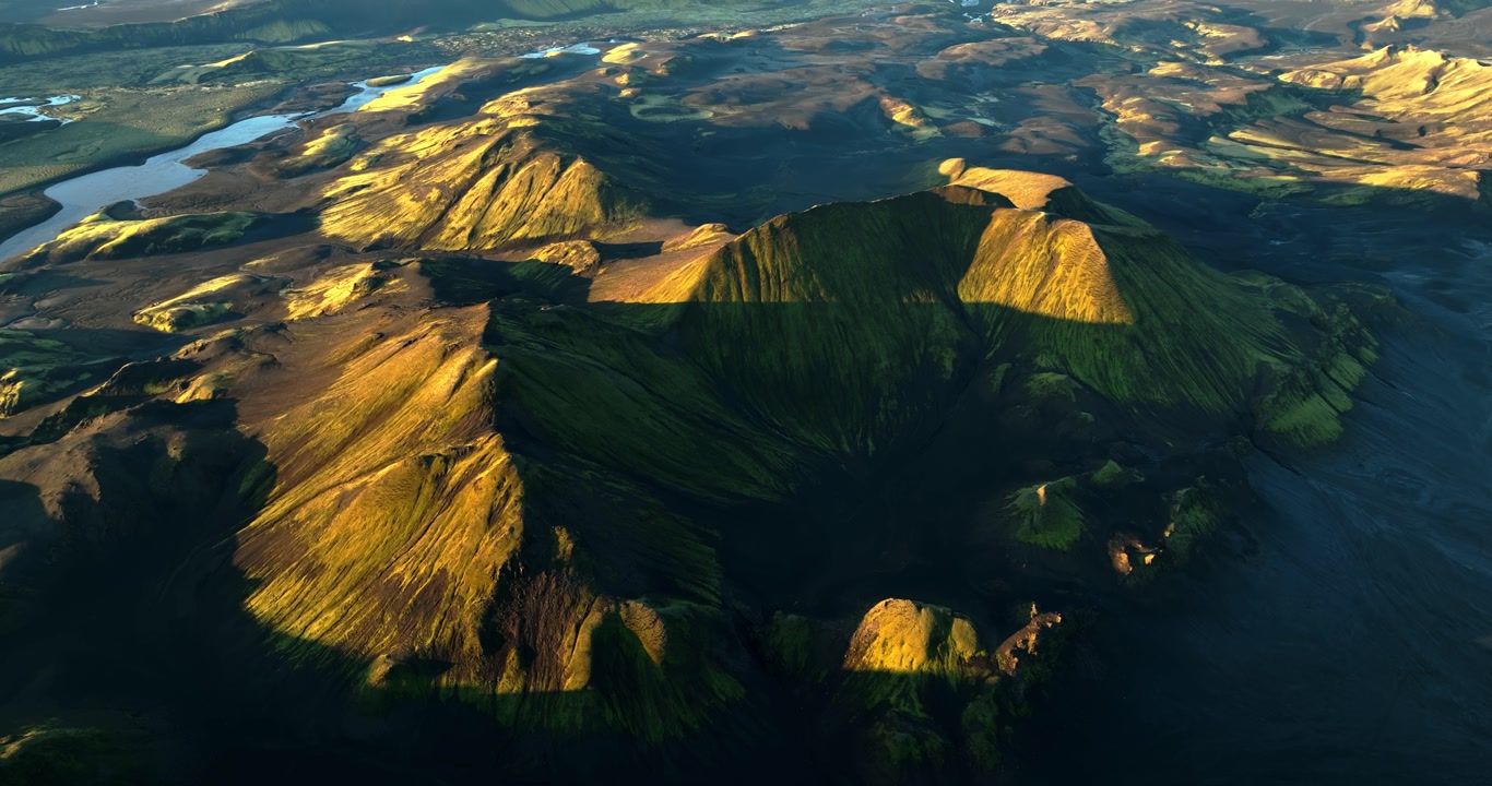 Drone flyover of a moss-covered volcanic crater in Iceland highlands, golden-hour light and deep shadows across ash slopes, with rivers and lava plains in the distance.