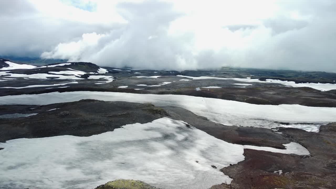 Aerial drone footage flying and hovering over cold and white snow covered Iceland volcanic dark and rocky landscapes during a cloudy day with large mountains visible in the background.
