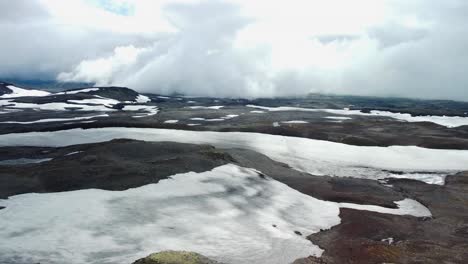 aerial drone footage flying and hovering over cold and white snow covered iceland volcanic dark and rocky landscapes during a cloudy day with large mountains visible in the background.