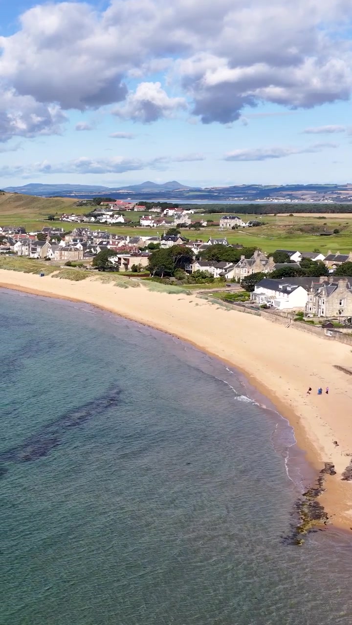 En drone svæver over Dundees kystlinje og afslører landsbyhuse, sandstrand, klart hav og blå himmel.