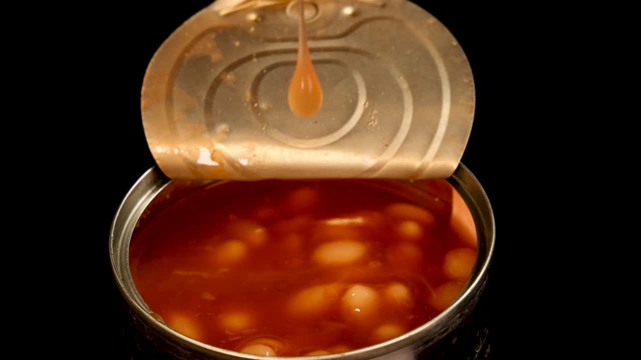 A metal spoon lifts baked beans in tomato sauce from an open can against a black background, with close-up, well-lit shots and smooth camera movement
