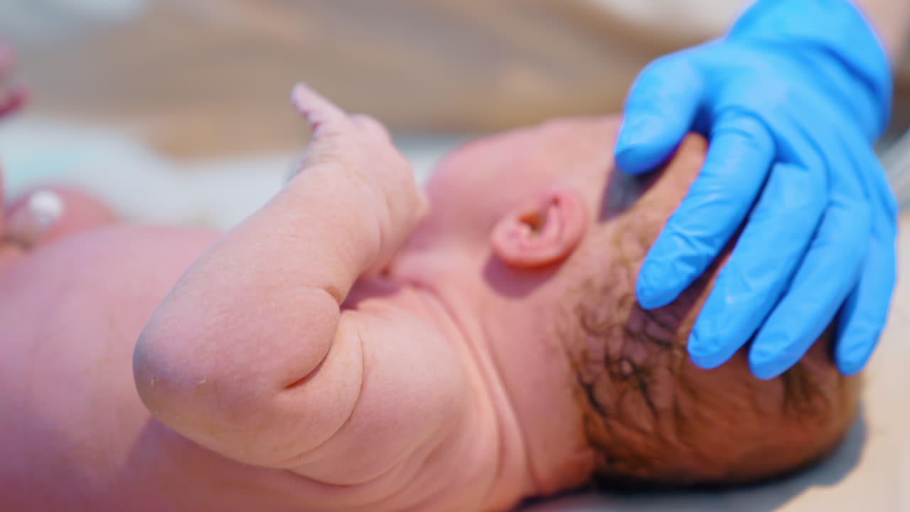 Hand in blue latex glove strokes the head of a newborn. Caucasian baby lies naked on the table. Close up.