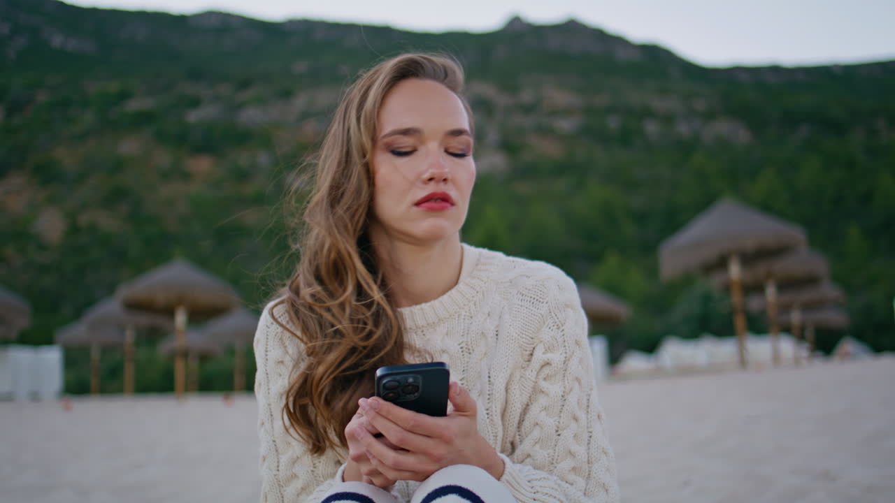 Confused woman watching cellphone sitting evening seashore alone closeup