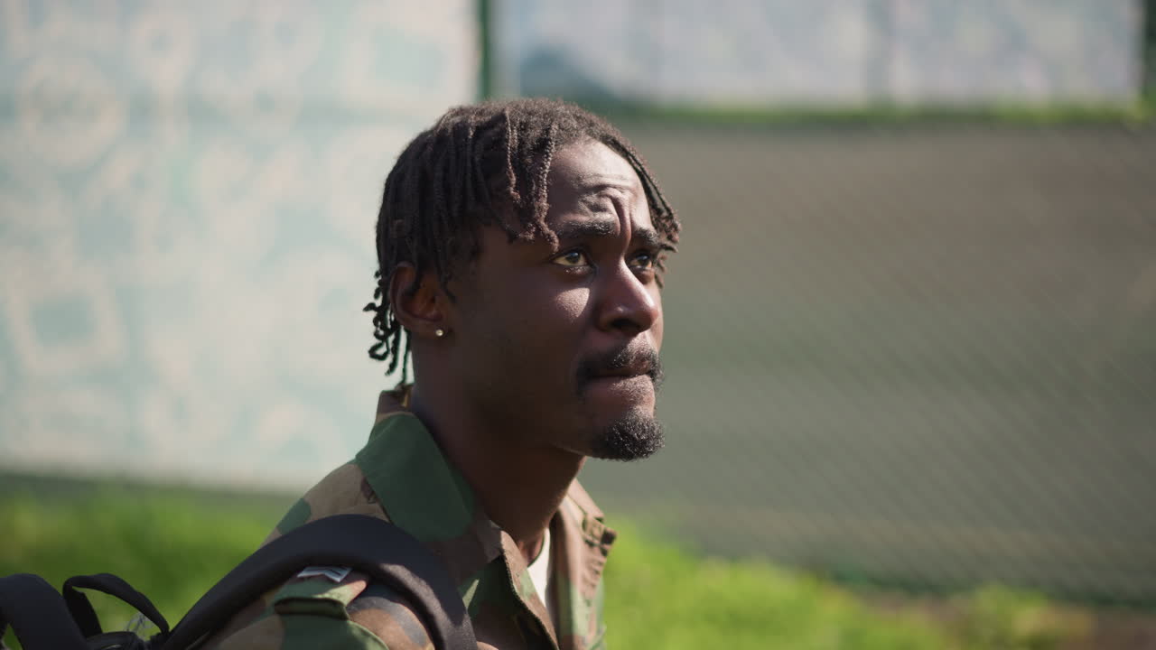 Black Soldier Profile With Backpack Looking Upward Near Fenced Court, Closeup Portrait Shows Dreadlocks, Thoughtful Expression, Camouflage Jacket, Gentle Sunlight, Introspective Urban Vibe