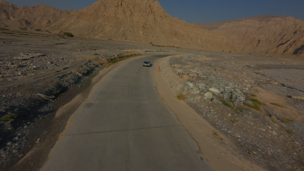 FPV drone chase through Hatta Desert following a car driving on a winding road. The drone catches up from behind, flies alongside the vehicle, and continues forward over the arid rocky landscape