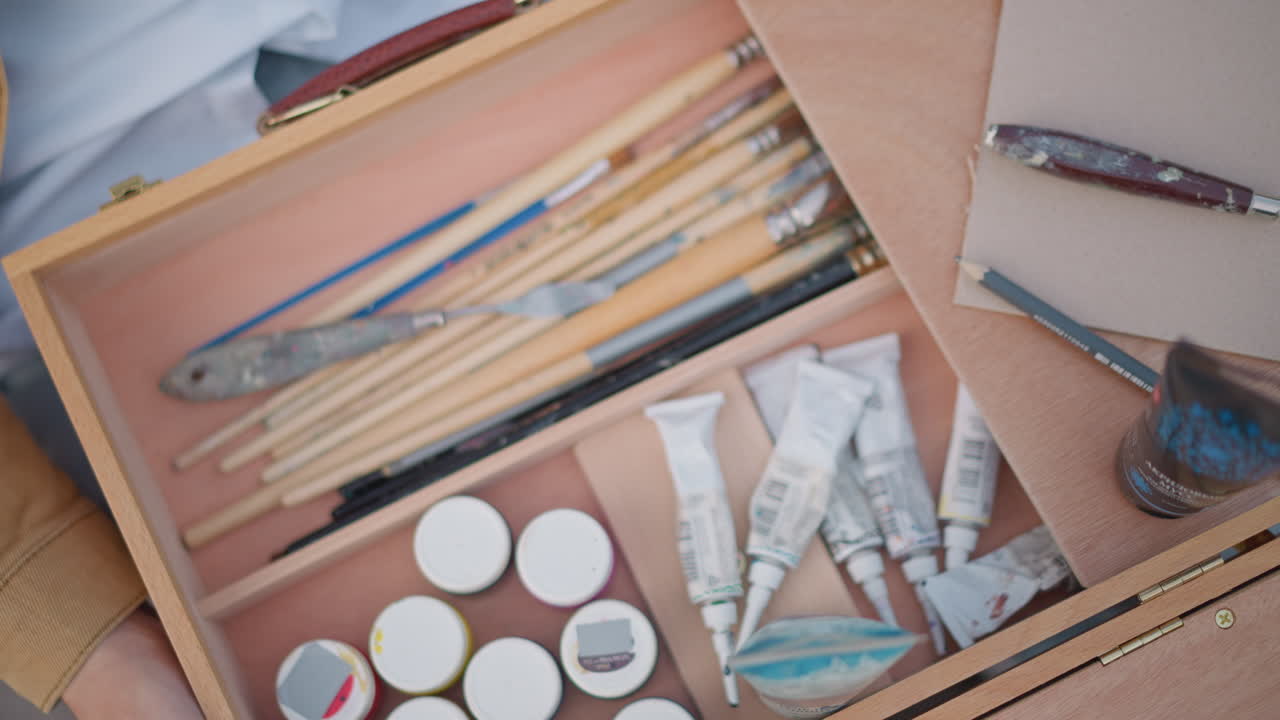 kit di pittura portatile da esterno, scatola di legno, pennelli allineati accanto a piccoli barattoli e tubetti spremuti, mani di artista che tengono il bordo della valigetta pronti per il disegno dal vero, spatola e setole visibili, urbano