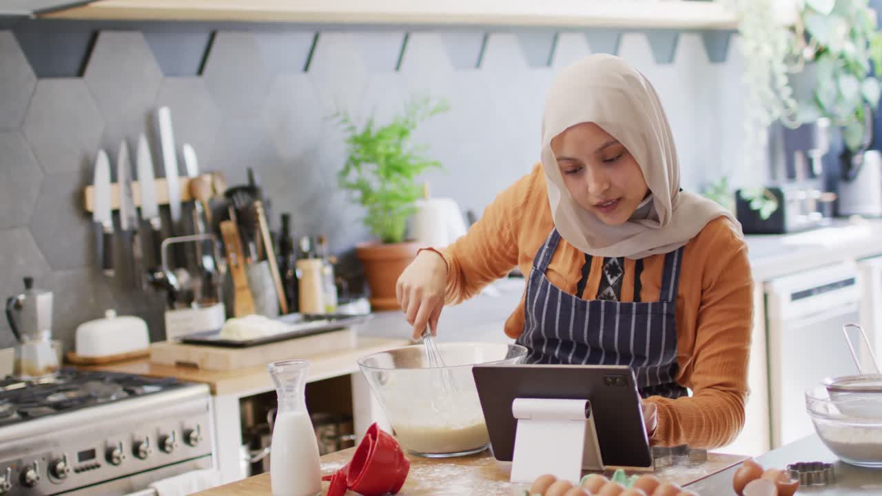 video de una feliz mujer biracial con hijab horneando en la cocina de su casa, usando la receta en una tableta