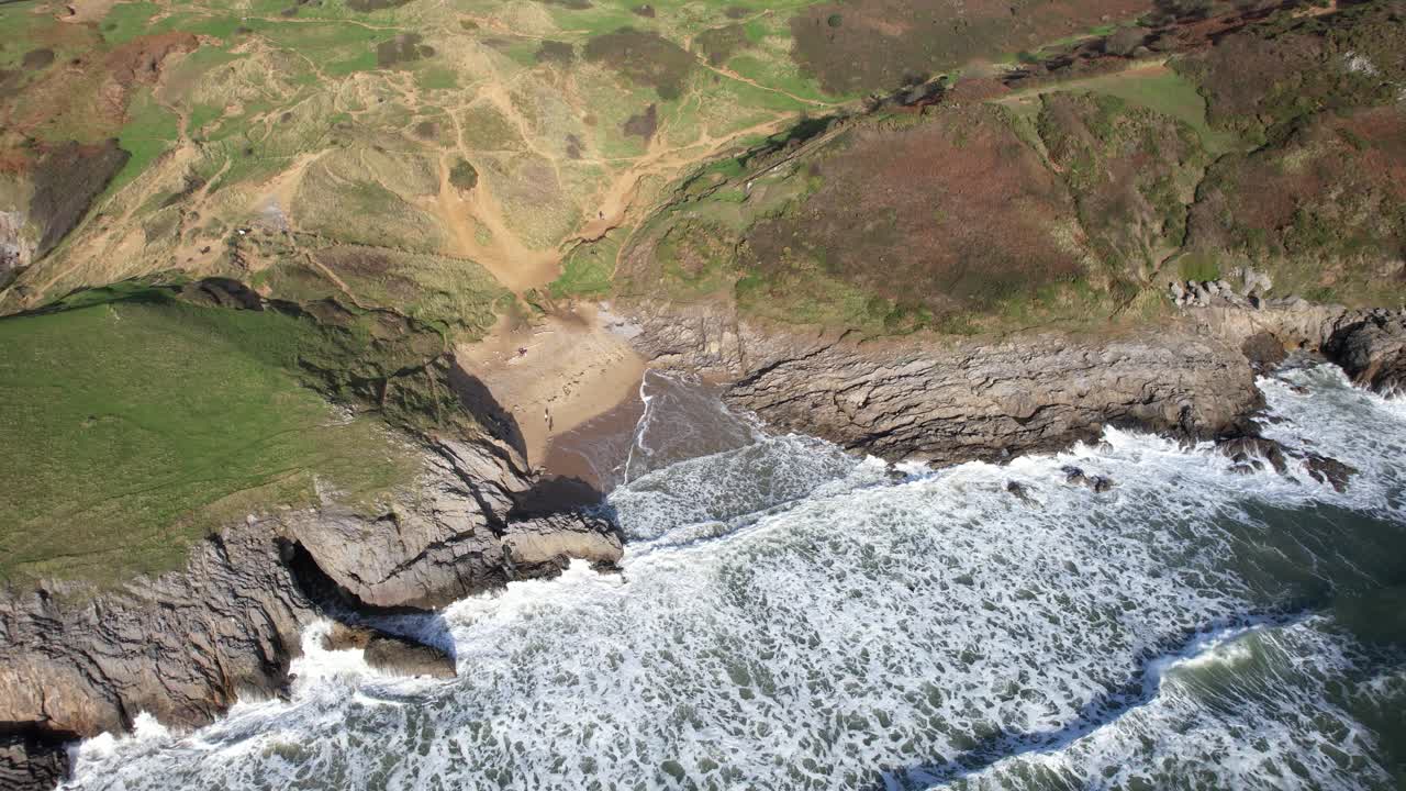 Circling view over Pobbles Bay with foaming waves rocks and coastal grasslands along the Gower Peninsula shoreline in Wales