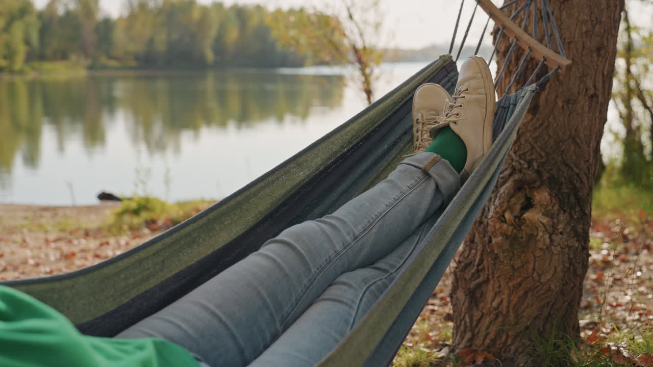 Serene female resting by water. Woman enjoying peaceful lakeside rest. Tranquil woman lounging on hammock by scenic lake. Calm woman in jeans soaking in peaceful lakeside autumn scenery
