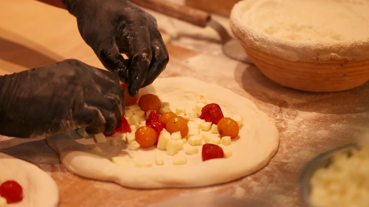 Primo piano di mani guantate che posizionano pomodorini freschi su impasto crudo per pizza con mozzarella.