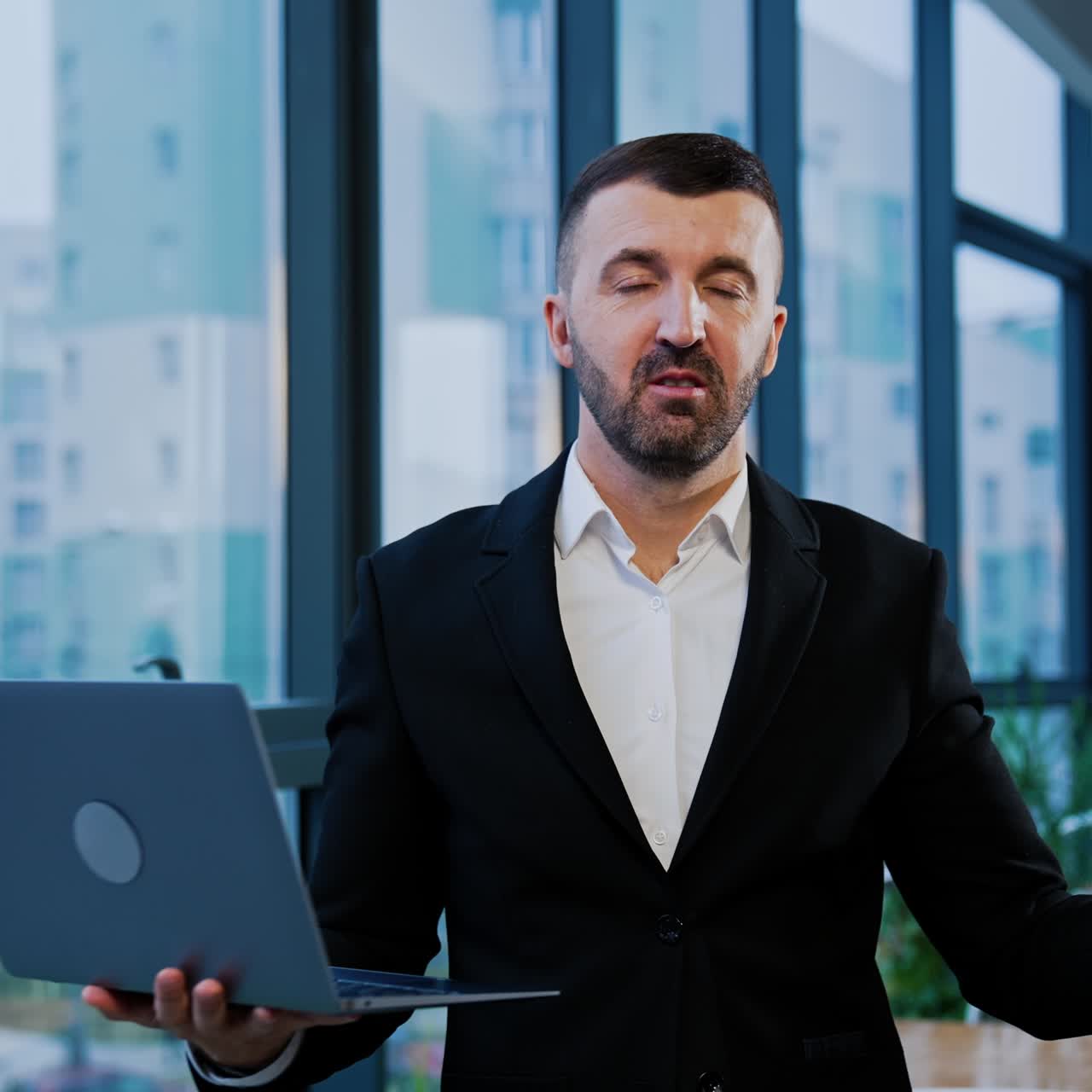 Smiling good-looking bearded businessman in suit stands near the window holding laptop. Man talks showing something with his hands