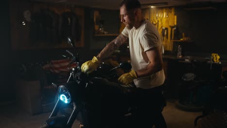 mecánico de tipo confiado en una camiseta blanca se pone guantes amarillos y se sienta en su bicicleta en el garaje del taller