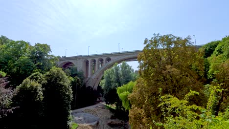 camera pans across lush green park toward a historic stone arch bridge under clear blue sky, capturing vibrant summer foliage and architectural details