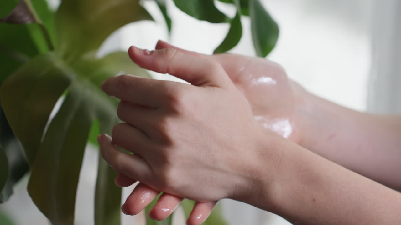 Light skin woman gently scrubbing oily palms together beside green leaf, hands glowing under natural soft light, emphasizing wellness, self-care, and calm spa-like atmosphere indoors