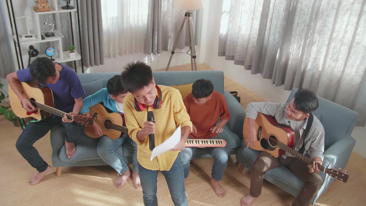 로크 음악을 연주하는 젊은 아시아 친구 그룹, 소년이 노래하고 종이에서 가사를 읽는 동안 그의 친구들은 집에서 소파에서 악기를 연주합니다.
