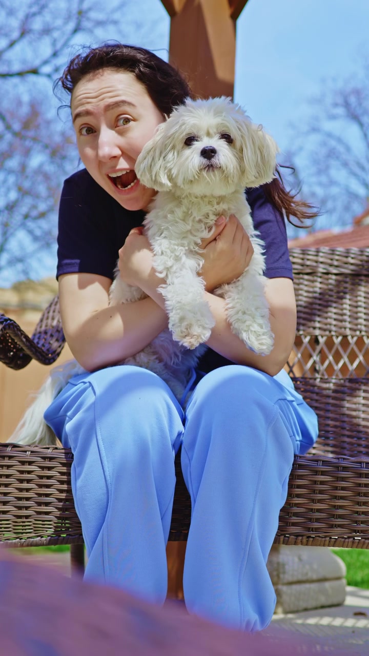 Woman sits on a bench and pets small white dog in sunny outdoor area while trees are in the background during daytime