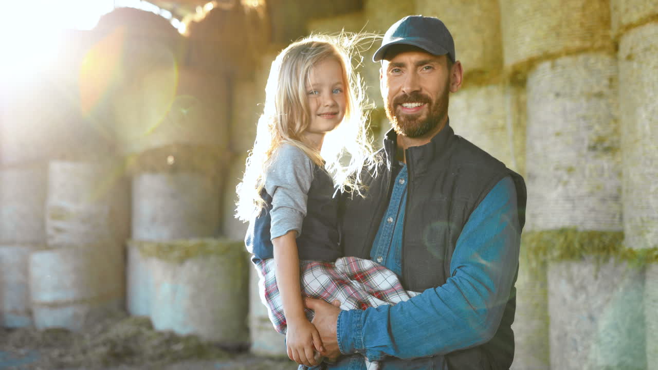 Portrait of Caucasian young father holding on hands his cute little daughter and smiling at camera in stable with hay stocks