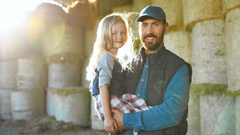 portrait of caucasian young father holding on hands his cute little daughter and smiling at camera in stable with hay stocks