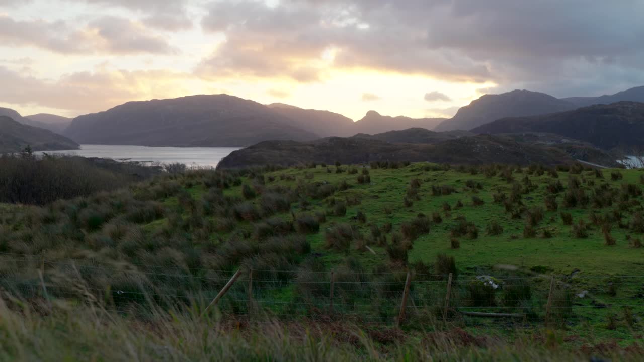 강한 바람이 전경의 풀과 하늘을 가로지르는 구름을 불고 스코틀랜드의 배경에 있는 산 뒤에 황금빛 태양이 지는 동안