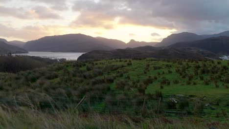 Un-Fuerte-Viento-Sopla-Hierba-En-Primer-Plano-Y-Nubes-En-El-Cielo-Mientras-Un-Sol-Dorado-Se-Pone-Detrás-De-Las-Montañas-En-El-Fondo-En-Escocia