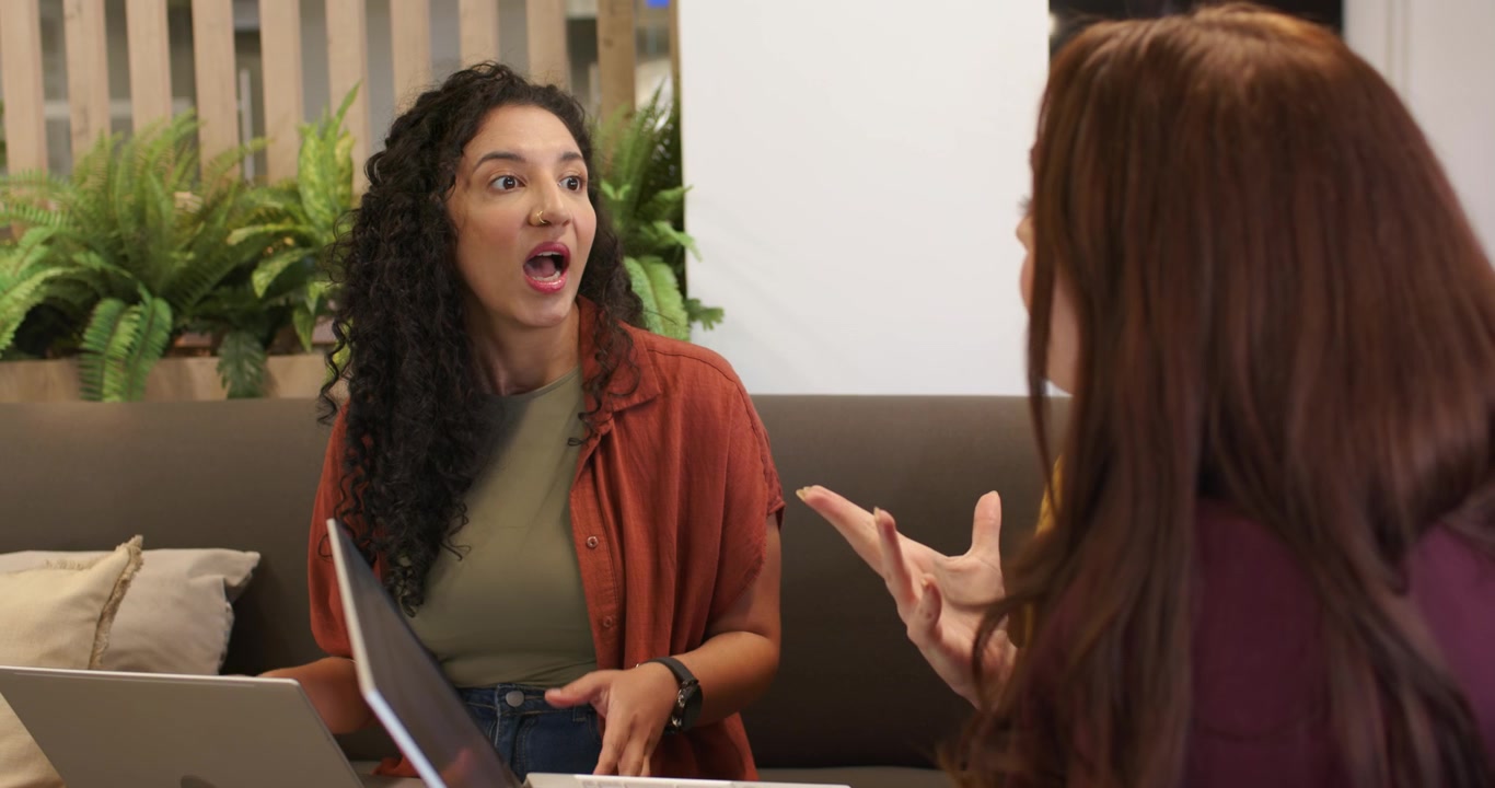 Colegas femeninas conversando, la oradora de la izquierda comienza a hablar, gesticulando en el sofá con ordenadores portátiles.
